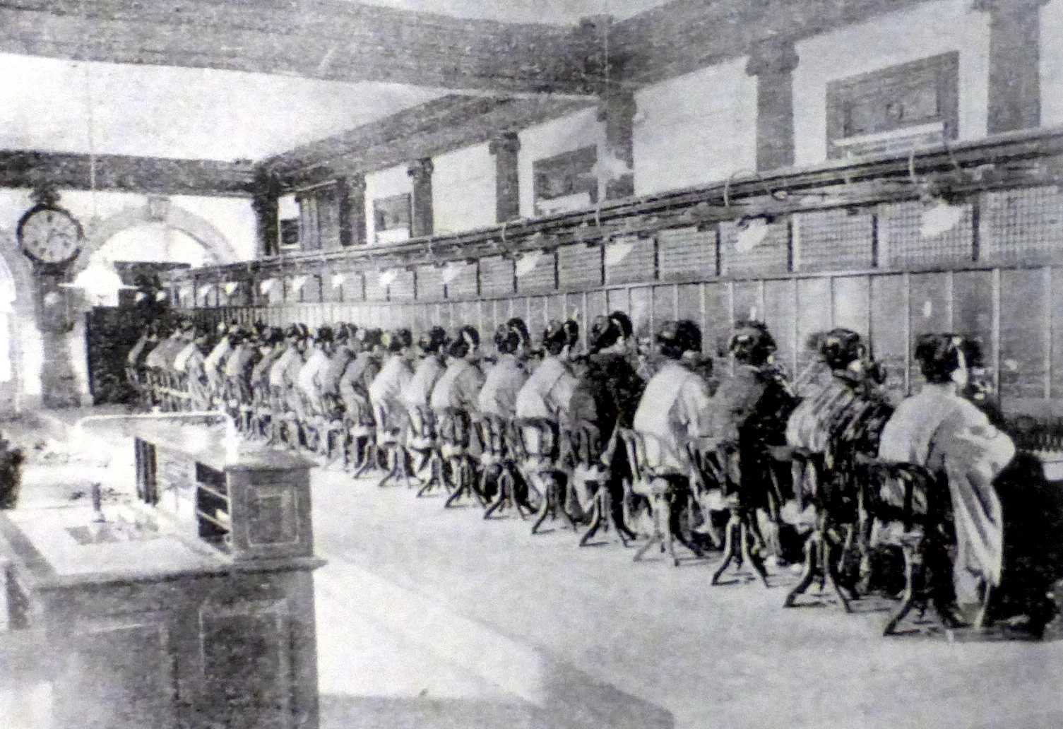 A long room of telephone operators in Japan, 1902, seated in a row along the switchboard